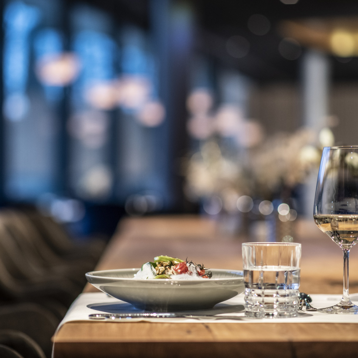 Ein elegant gedeckter Tisch in einem stilvollen Restaurant. Im Vordergrund steht ein appetitlich angerichteter Teller, daneben ein Glas Wasser und zwei Weingläser. Sanfte Beleuchtung und moderne Einri