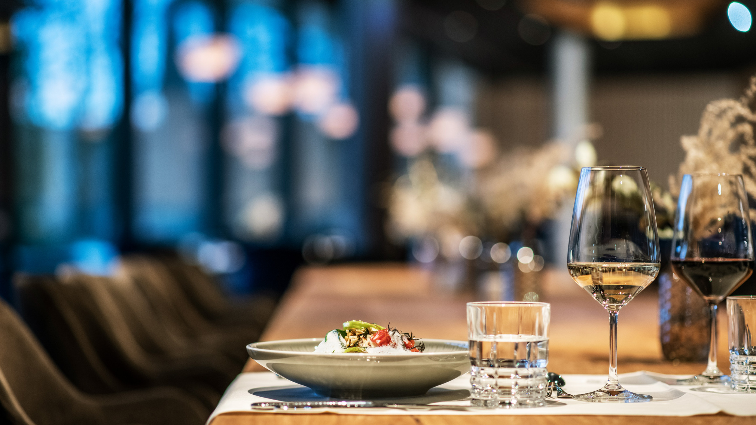 Ein eleganter Restauranttisch in warmem Licht, mit einem gefüllten, grauen Teller, Gläsern für Wein und Wasser sowie sanften, verschwommenen Hintergrundelementen und stilvollen Sitzmöbeln.