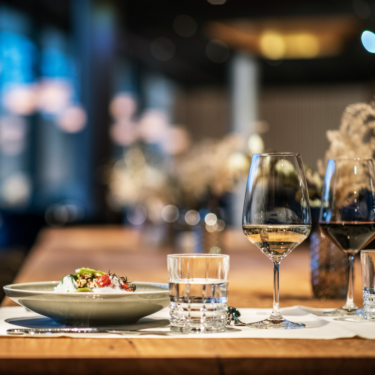 Ein eleganter Restauranttisch in warmem Licht, mit einem gefüllten, grauen Teller, Gläsern für Wein und Wasser sowie sanften, verschwommenen Hintergrundelementen und stilvollen Sitzmöbeln.