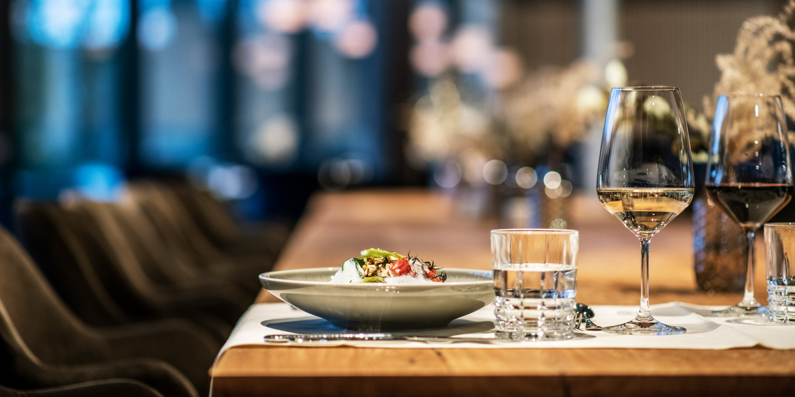 Ein eleganter Restauranttisch in warmem Licht, mit einem gefüllten, grauen Teller, Gläsern für Wein und Wasser sowie sanften, verschwommenen Hintergrundelementen und stilvollen Sitzmöbeln.