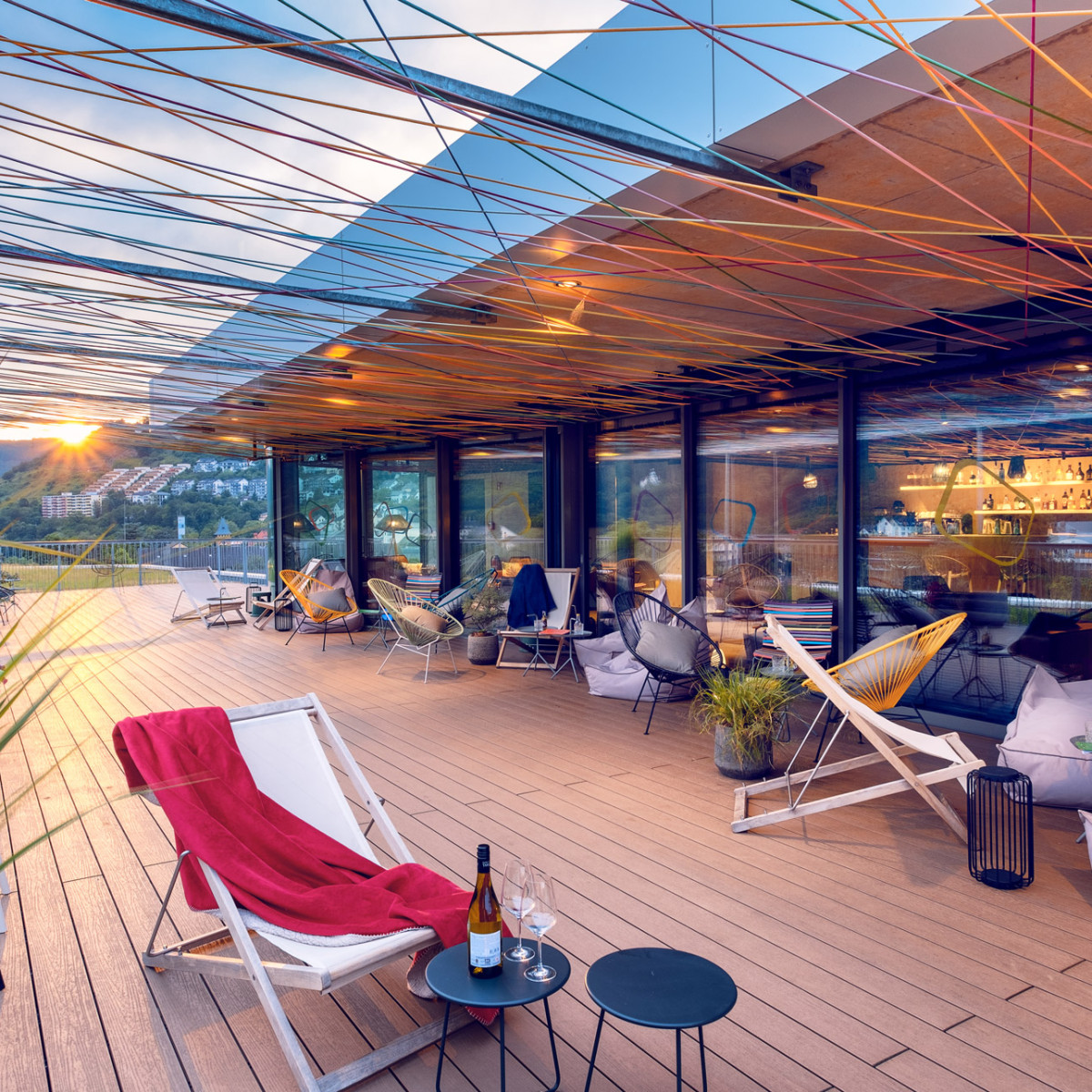 Gemütliche Terrasse mit modernem Design, geschmückt mit bunten Fäden. Sonnenuntergang im Hintergrund, Loungemöbel, Pflanzen und Getränke auf kleinen Tischen laden zum Entspannen ein.