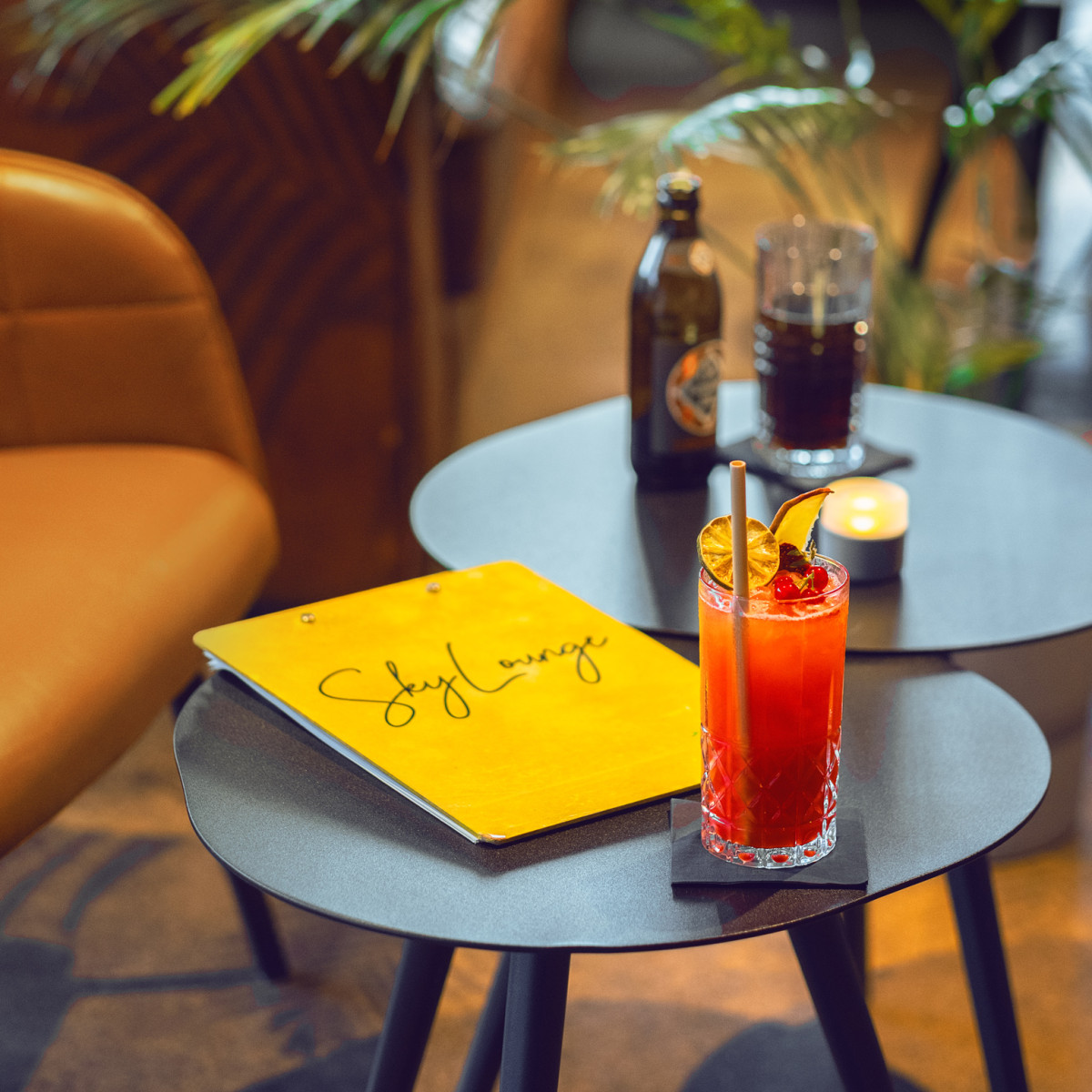 Ein stilvoller Tisch mit einem bunten Cocktail, einer Getränkekarte in Gelb mit der Aufschrift "Sky Lounge", einem Glas Cola und einer Kerze, umgeben von Pflanzen und einem gemütlichen Ambiente.