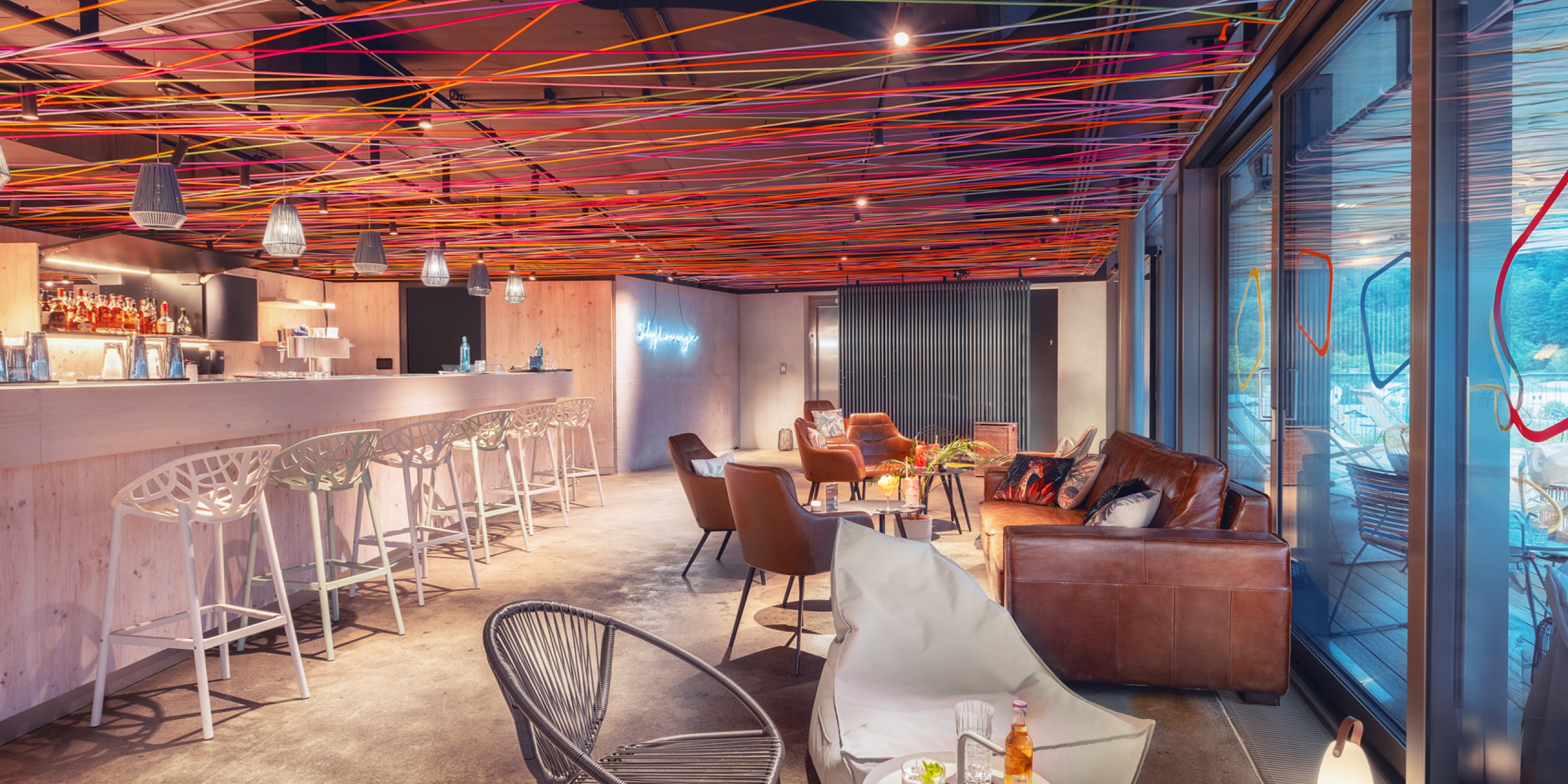 Moderner Lounge-Bereich mit bunten, geflochtenen Linien an der Decke. Eine stilvolle Bar mit Hochtischen, gemütlichen Sesseln und einer einladenden Atmosphäre. Große Fenster mit Blick nach draußen.