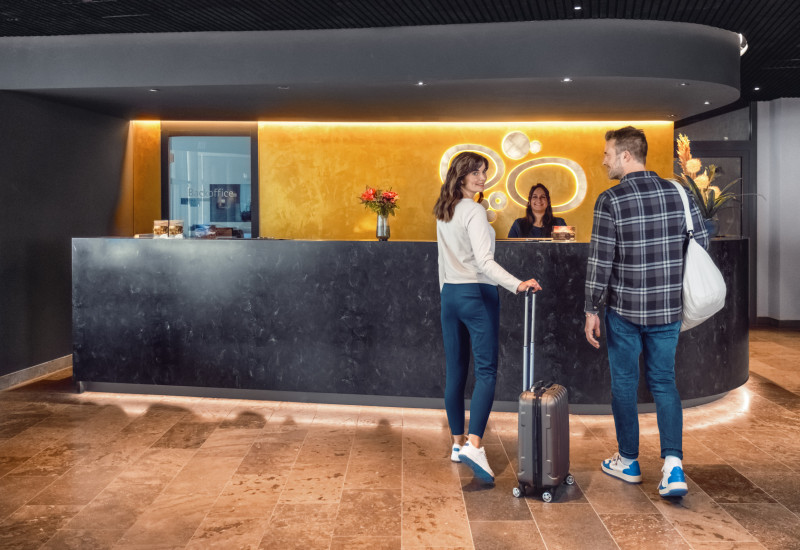 Moderne Hotellobby mit einem Empfangstresen, an dem eine Frau mit einem Koffer und ein Mann im Gespräch mit einer Hotelangestellten stehen. Blumen dekorieren den Tresen.