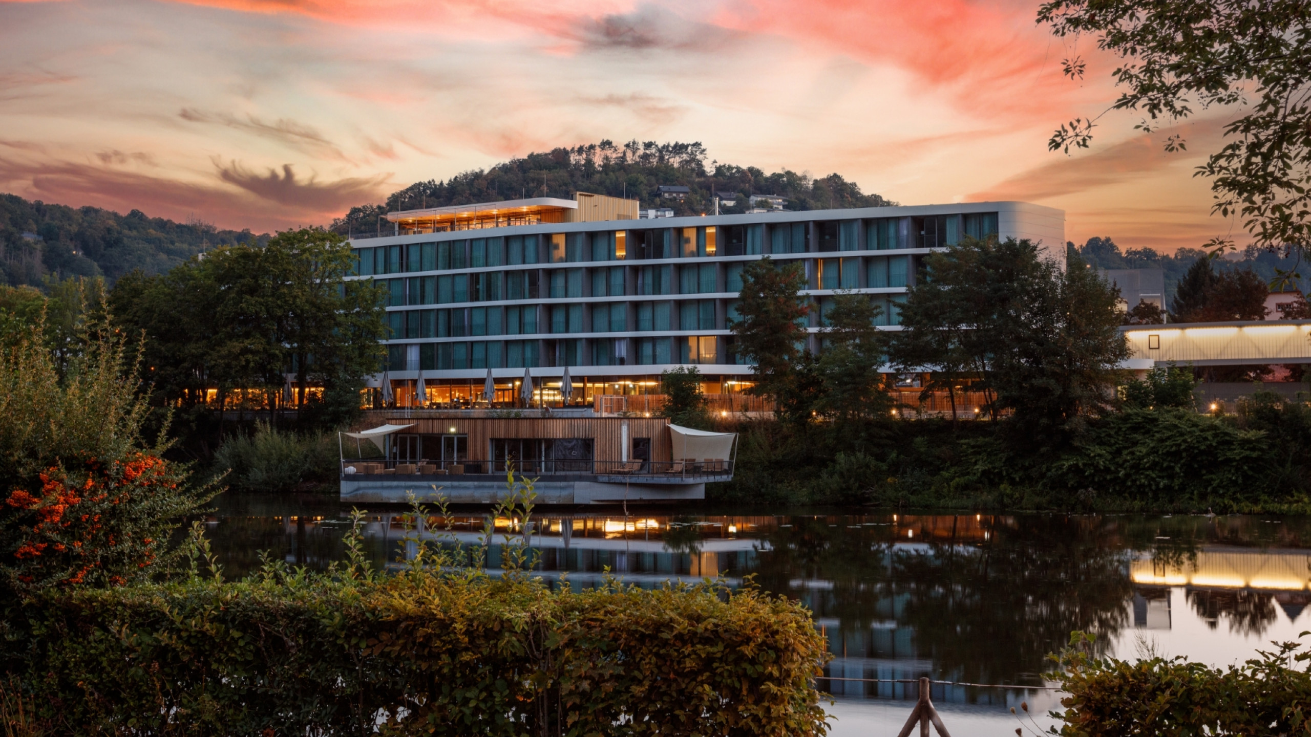 Modernes Hotel am Ufer eines ruhigen Sees, umgeben von Bäumen und grünen Sträuchern. Im Hintergrund ein sanft beleuchteter Hügel und ein farbenfroher Sonnenuntergang am Himmel.