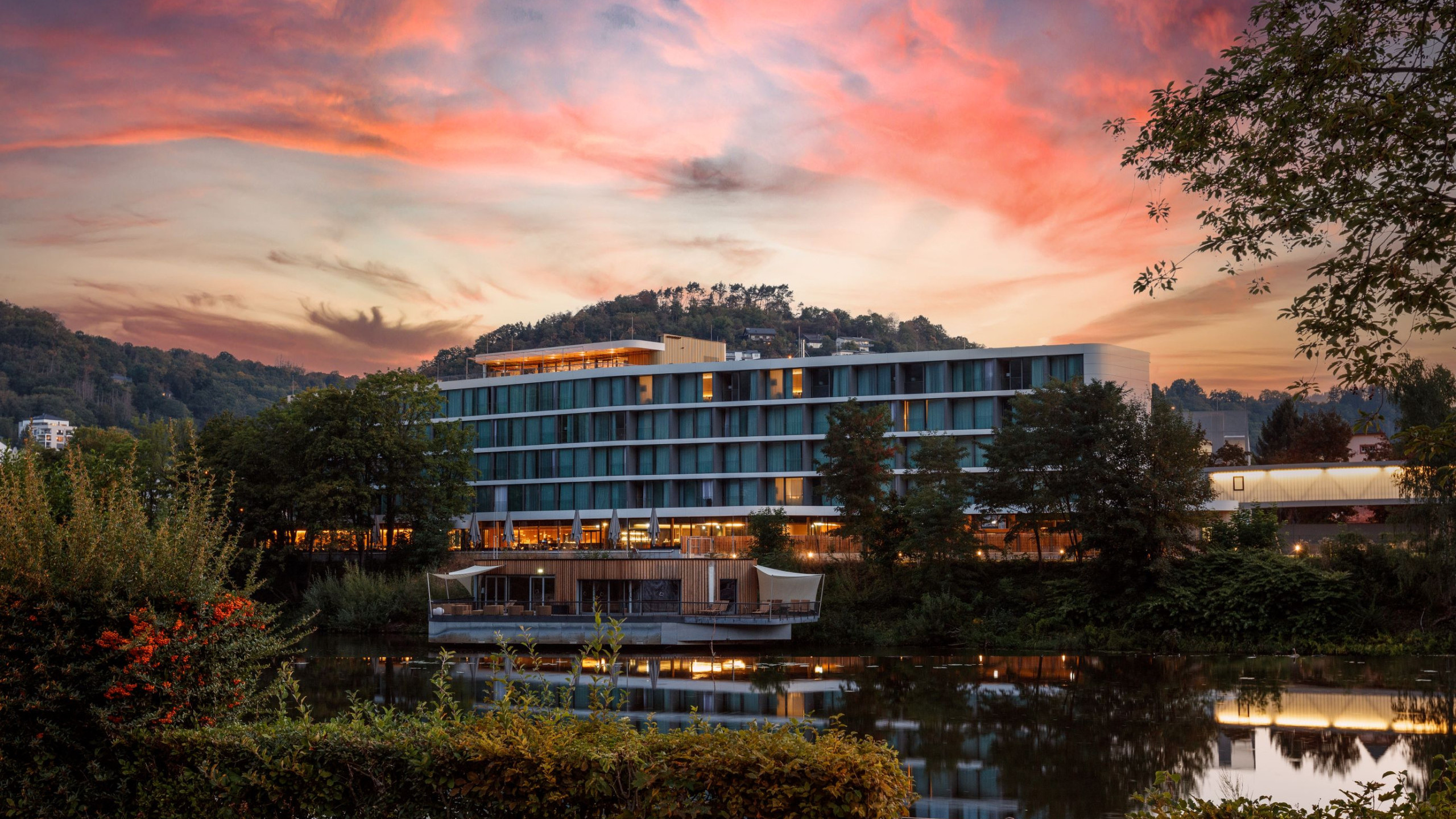 Moderne Hotelarchitektur am Ufer eines ruhigen Gewässers, umgeben von Bäumen und Hügeln. Ein lebendiger Sonnenuntergang mit pinken und orangefarbenen Wolken spiegelt sich im Wasser.