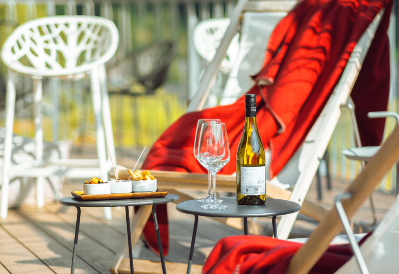 Gemütliche Terrasse mit einem gefüllten Weinglas, einer Flasche Wein und köstlichen Snacks auf einem Tisch. Ein rotes Plaid liegt über einem Liegestuhl, umgeben von modernen Gartenstühlen.