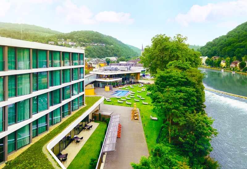 Moderne Hotelanlage am Flussufer, umgeben von grüner Landschaft. Die Architektur zeigt große Fenster und Terrassen. Entspannungsbereiche mit Liegen und einem kleinen Pool sind sichtbar.