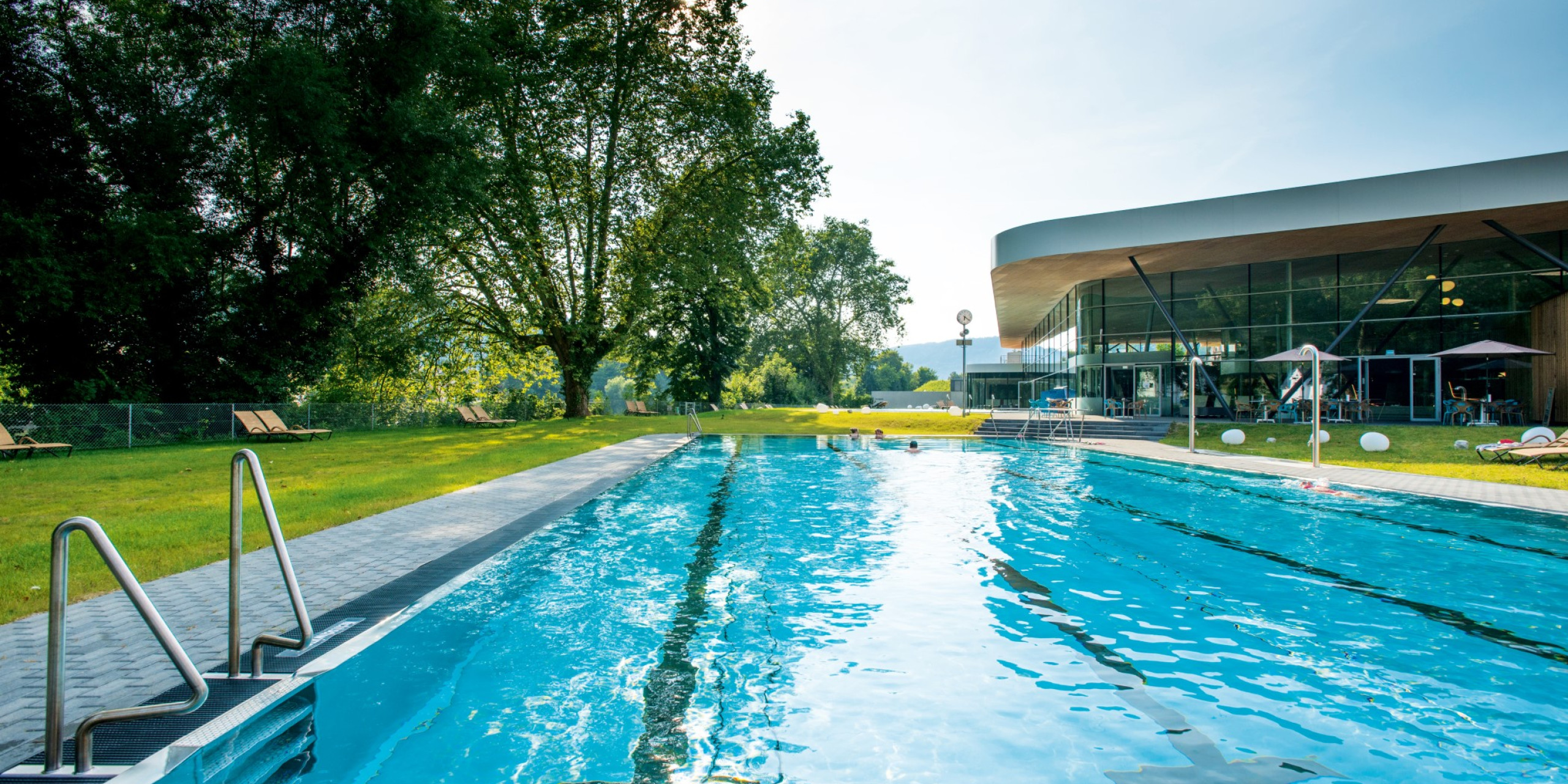 Ein einladender Schwimmbeckenbereich, umgeben von grünen Rasenflächen und Bäumen. Im Hintergrund ist ein modernes Gebäude mit großen Fenstern sichtbar, Sonnenlicht reflektiert auf dem Wasser.