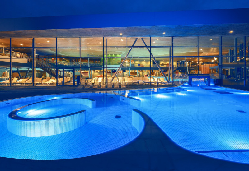 Beleuchteter Pool mit geschwungener Form, umgeben von modernen Liegen. Im Hintergrund sind große Fenster, die eine stilvolle Innenarchitektur und weitere Einrichtungen zeigen. Abendliche Atmosphäre.