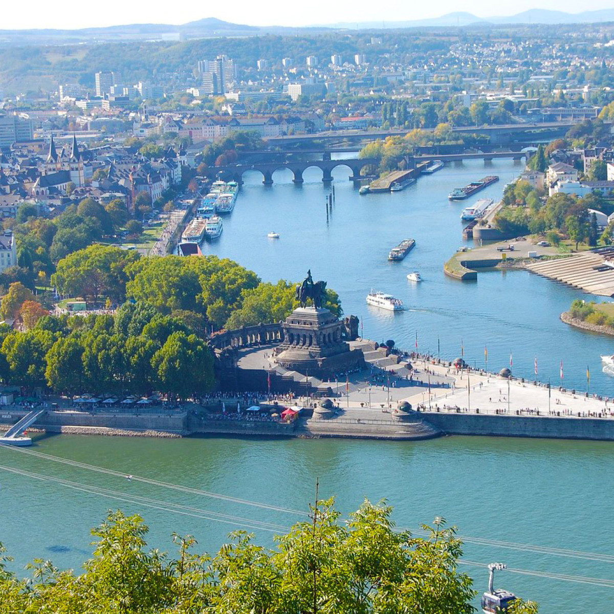 Panoramablick über das Rheintal mit dem Deutschen Eck, wo Mosel und Rhein zusammenfließen. Grüne Bäume umrahmen die Ufer, zahlreiche Boote sind auf dem Wasser unterwegs, und die Stadt ist sichtbar.