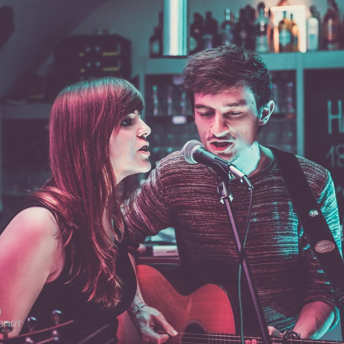 Zwei Musiker stehen auf einer kleinen Bühne in einer gemütlichen Bar. Die Frau singt in ein Mikrofon, während der Mann mit einer Akustikgitarre spielt. Bunte Lichter schaffen eine entspannte Atmosphär