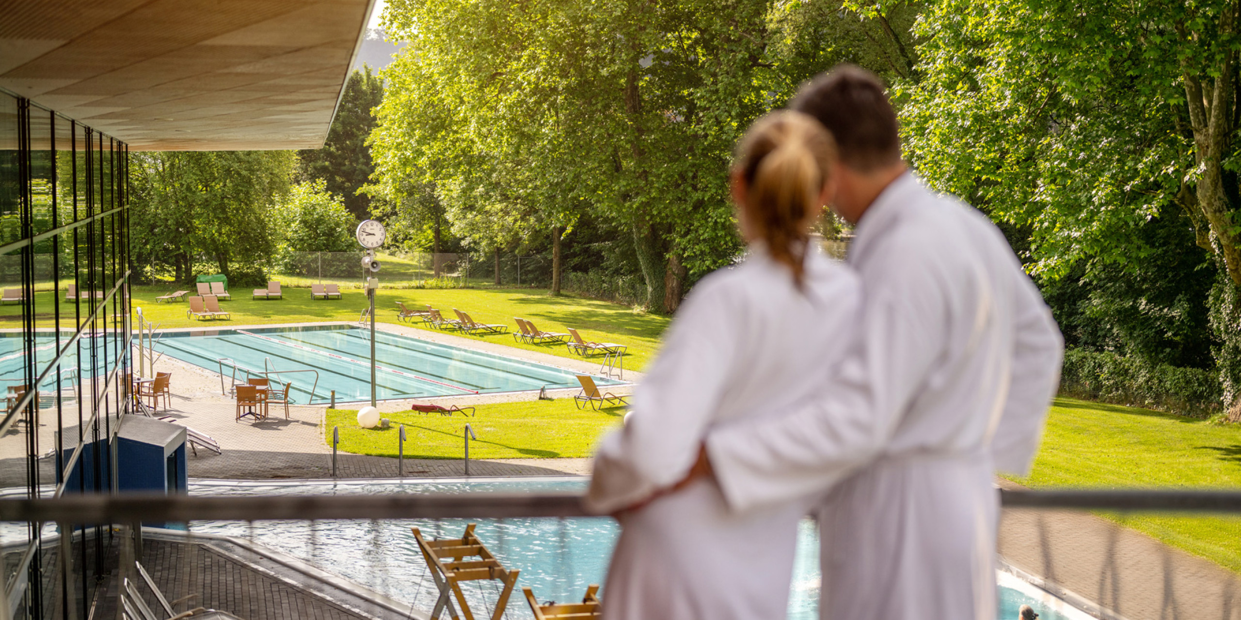 Ein Paar in weißen Bademänteln steht auf einem Balkon und blickt auf einen Außenpool, umgeben von grünem Rasen und Bäumen. Entspannte Atmosphäre mit Sonnenstrahlen.