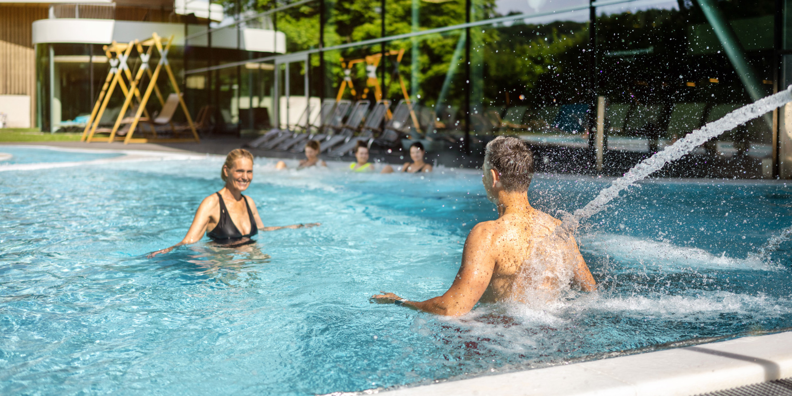 Zwei Personen genießen einen sonnigen Tag im Pool. Eine Frau in einem schwarzen Bikini lacht, während ein Mann im Wasser spritzt. Im Hintergrund sind Liegen und eine moderne Architektur zu sehen.