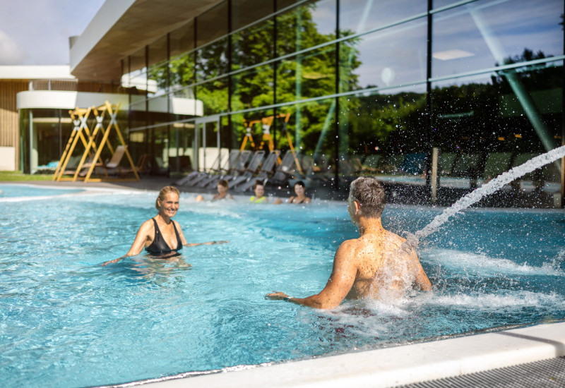 Zwei Personen genießen einen sonnigen Tag im Pool. Eine Frau in einem schwarzen Bikini lacht, während ein Mann im Wasser spritzt. Im Hintergrund sind Liegen und eine moderne Architektur zu sehen.