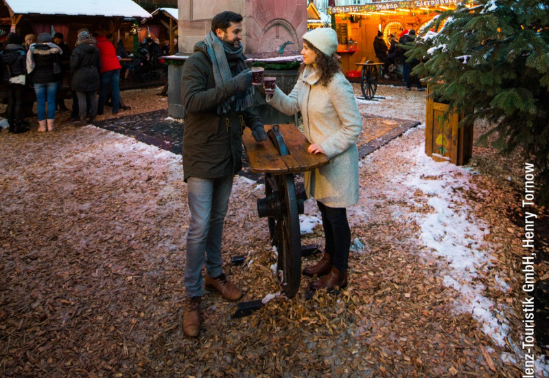 Ein Paar stößt mit Glühwein an, umgeben von festlicher Weihnachtsmarkt-Atmosphäre. Beleuchtete Stände und ein dekorierter Weihnachtsbaum im Hintergrund, Schnee auf dem Boden.