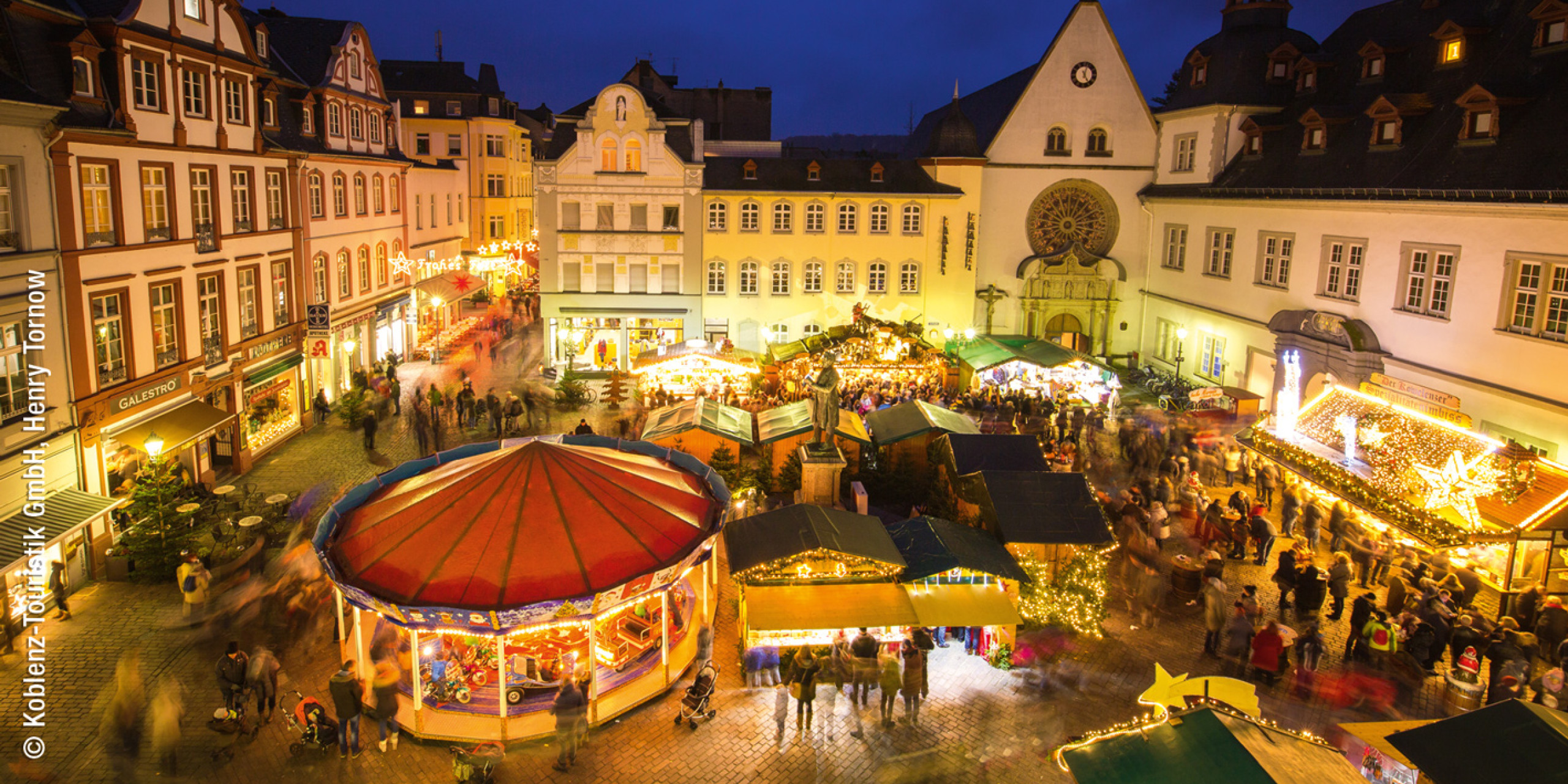 Beleuchteter Weihnachtsmarkt bei Dämmerung, umgeben von historischen Gebäuden. Buden mit festlichen Lichtern, ein Karussell und Menschen, die die festliche Atmosphäre genießen.