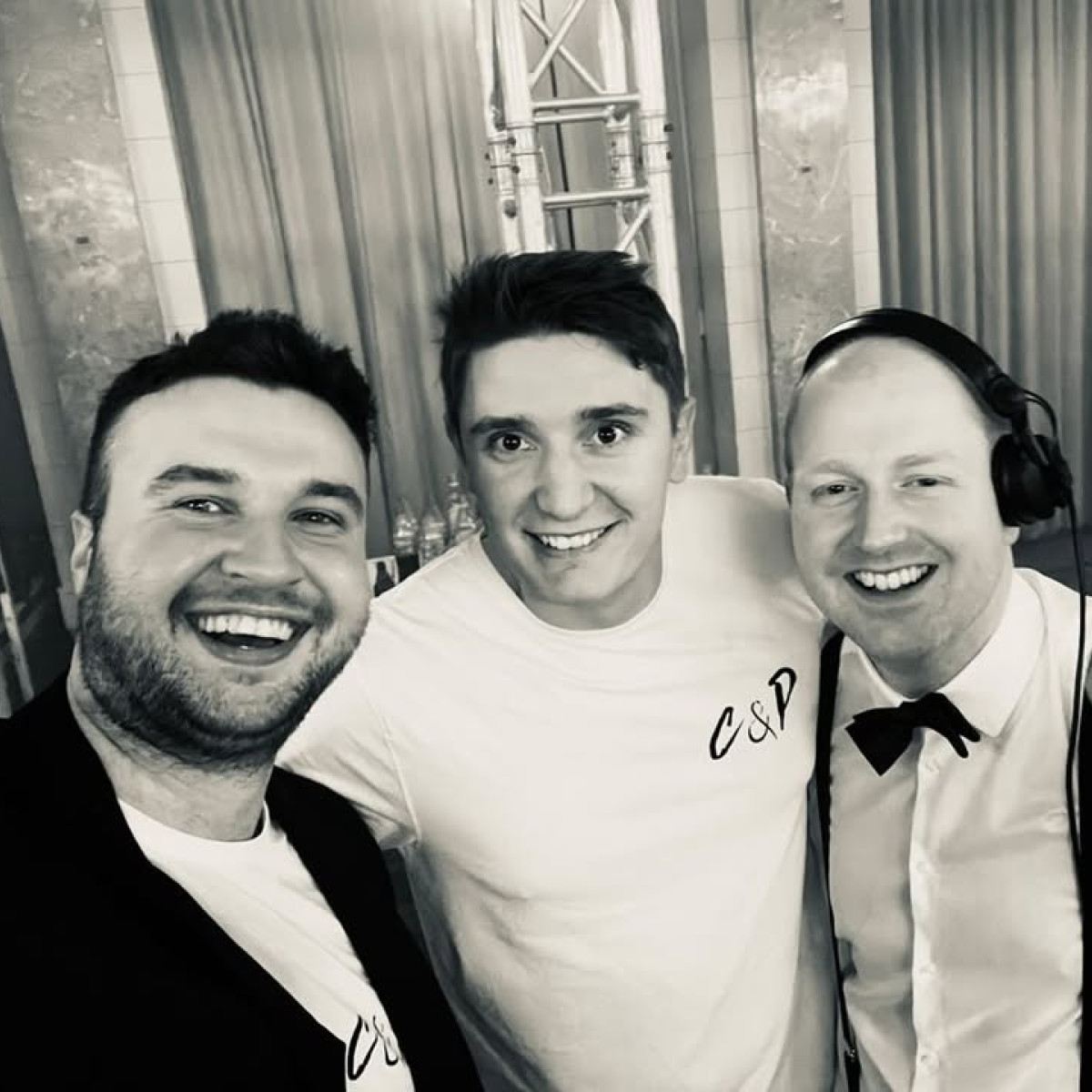 Drei Männer lächeln in die Kamera, stehen eng beieinander. Der linke trägt einen dunklen Blazer, der mittlere ein T-Shirt mit „C&P“ und der rechte eine Fliege mit Kopfhörern. Hintergrund ist elegant g