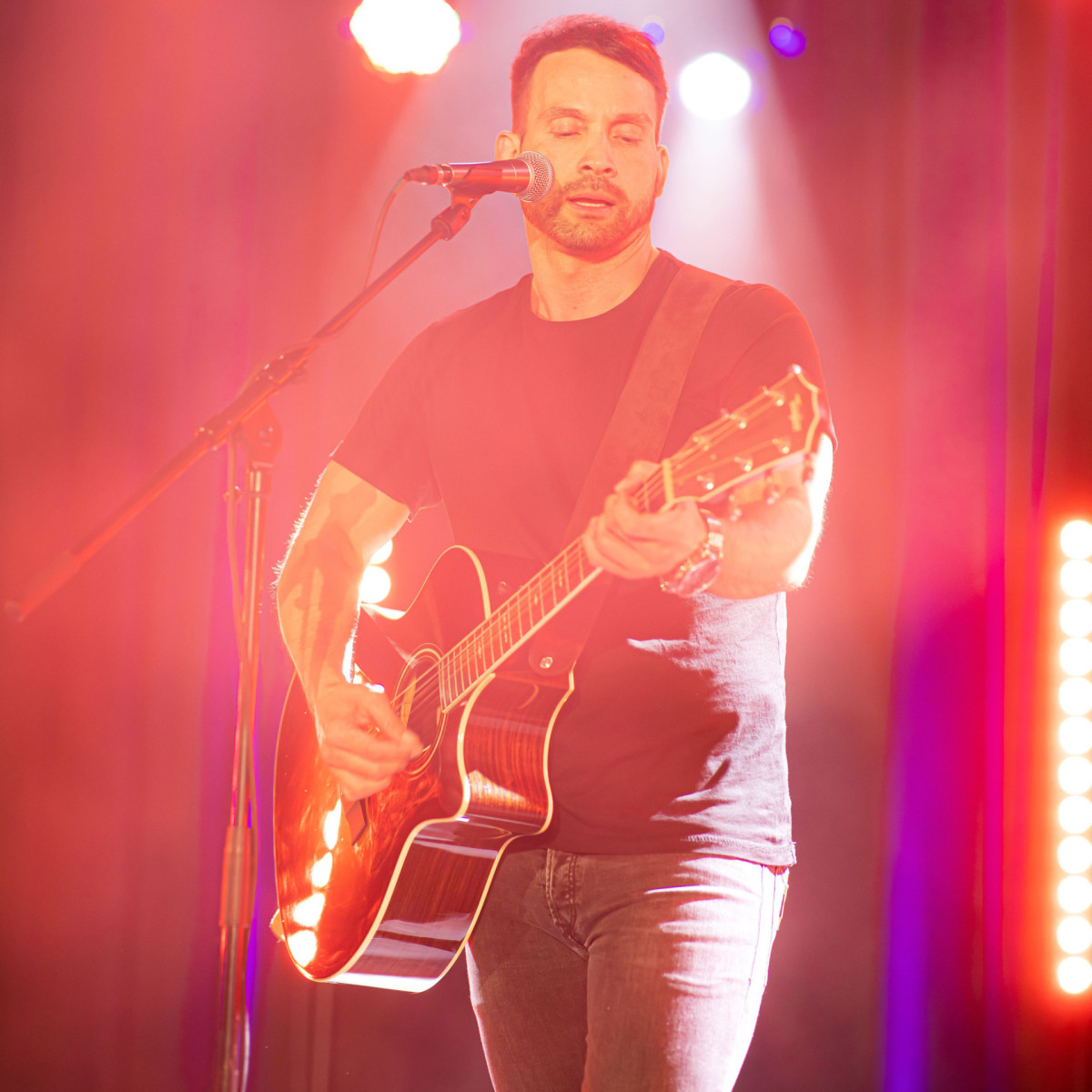 Ein Mann mit Bart spielt auf einer Akustikgitarre auf der Bühne. Er trägt ein schwarzes T-Shirt und Jeans, umgeben von bunten Lichtern und Rauch, während er in ein Mikrofon singt.
