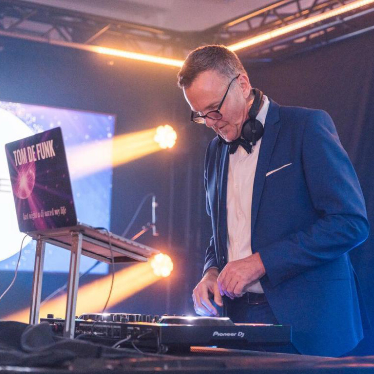 Ein DJ in einem eleganten Anzug mit Krawatte steht vor einem DJ-Setup und arbeitet konzentriert an den Turntables. Im Hintergrund sind bunte Lichtstrahlen zu sehen, die eine festliche Atmosphäre schaf