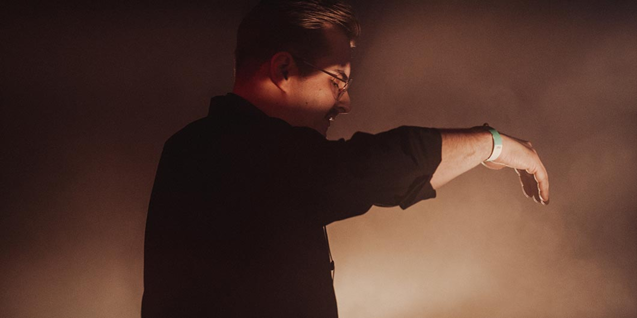 Eine silhouettenhafte Person in einem schwarzen Hemd steht vor einem nebligen, schwach beleuchteten Hintergrund. Die Hand ist angewinkelt, als ob sie mit der Umgebung interagiert.
