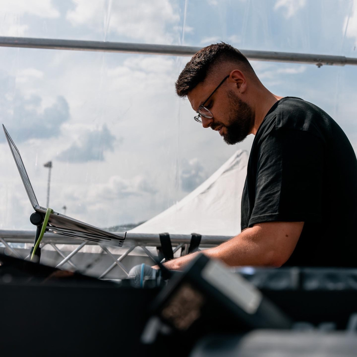 Ein DJ steht konzentriert an einem Mischpult unter einem durchsichtigen Zelt. Hinter ihm sind Wolken am Himmel sichtbar. Er trägt eine schwarze T-Shirt und eine Brille.